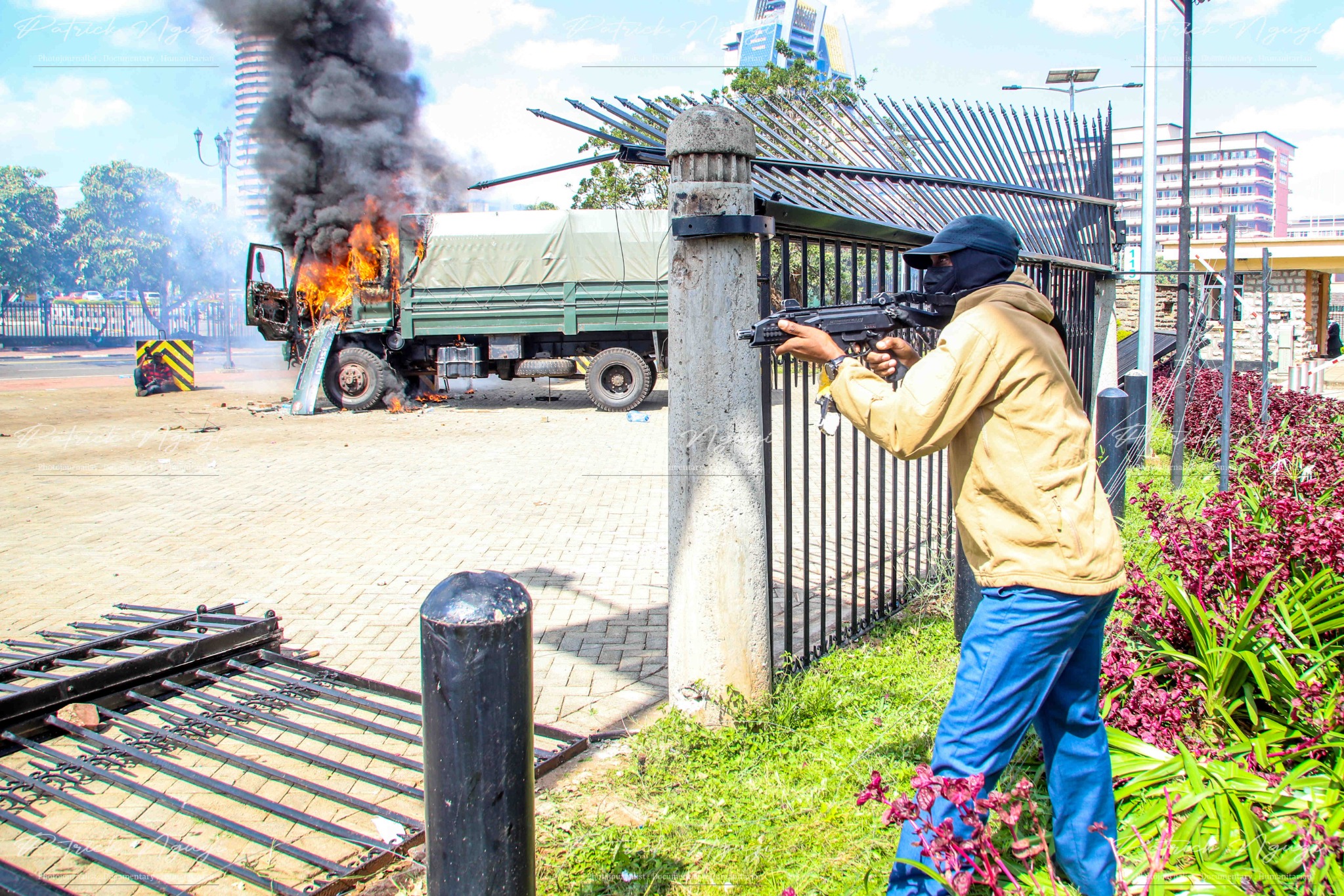 A masked man in a beige hoodie and blue trousers aims a firearm from behind a fence near the gates of Parliament in Nairobi. In the background, a GSU truck is engulfed in flames, thick black smoke rising into the sky. The surrounding area shows signs of destruction, with a fallen gate and scattered debris. The tension in the air reflects the intensity of the protests.