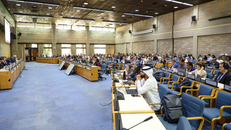 A wide conference hall at the United Nations Office in Nairobi filled with delegates seated at long rows of desks, many working on laptops and documents. At the front, a panel of speakers sits beneath large projection screens and a UNEP backdrop. Participants from various stakeholder groups—including scientific, local government, and civil society representatives—are engaged in drafting and amending environmental governance policies during UNEA-7.