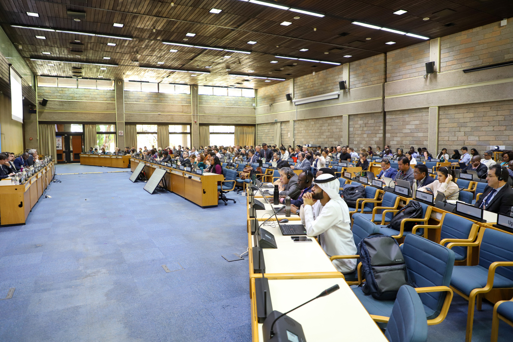 A wide conference hall at the United Nations Office in Nairobi filled with delegates seated at long rows of desks, many working on laptops and documents. At the front, a panel of speakers sits beneath large projection screens and a UNEP backdrop. Participants from various stakeholder groups—including scientific, local government, and civil society representatives—are engaged in drafting and amending environmental governance policies during UNEA-7.
