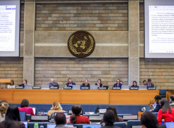A wide conference hall at the United Nations Office in Nairobi filled with delegates seated at long rows of desks, many working on laptops and documents. At the front, a panel of speakers sits beneath large projection screens and a UNEP backdrop. Participants from various stakeholder groups—including scientific, local government, and civil society representatives—are engaged in drafting and amending environmental governance policies during UNEA-7.
