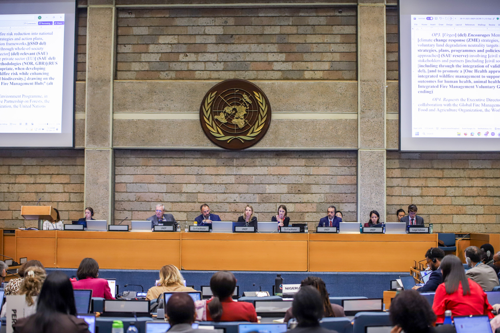 A wide conference hall at the United Nations Office in Nairobi filled with delegates seated at long rows of desks, many working on laptops and documents. At the front, a panel of speakers sits beneath large projection screens and a UNEP backdrop. Participants from various stakeholder groups—including scientific, local government, and civil society representatives—are engaged in drafting and amending environmental governance policies during UNEA-7.
